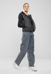 Schwarze gesteppte Weste mit Kapuze, Reißverschluss und Seitentaschen, kombiniert mit lockeren hellblauen Jeans und weißen Turnschuhen.