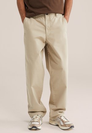 Personne portant un pantalon beige ample et des baskets crème et marron, debout les mains dans les poches devant un fond uni.