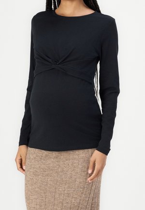 Kobieta w ciąży ubrana w czarną bluzkę z długim rękawem z przeplatanym węzłem i beżową prążkowaną dzianinową spódnicę, stojąca na białym tle.