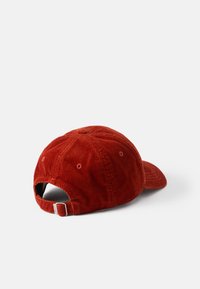 Punainen corduroy-baseball-lippis, jossa on pyöreä päällinen, kaareva lippa ja säädettävä kangasnauha takana. Näkyvä uritettu pinta.