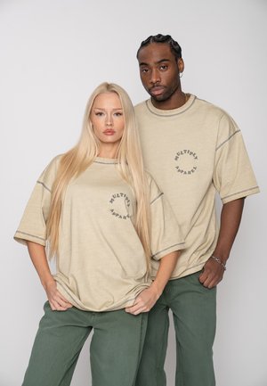 A man and woman wearing matching beige oversized t-shirts with "MULTIPLY APPAREL" text and green pants, standing closely against a plain background.