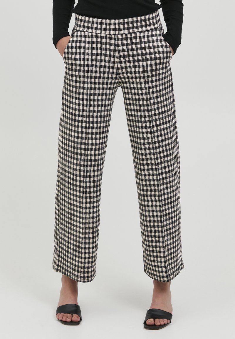 Pantalon large à motifs vichy noir et blanc avec poches latérales, taille haute et texture lisse. Porté avec des sandales plates noires.