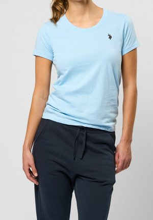 Kobieta ubrana w jasnoniebieską koszulkę z krótkim rękawem z małym logo konia oraz ciemnogranatowe spodnie z troczkami na tle jednolitego tła.