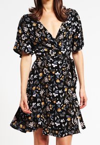 Mujer vestida con un vestido envolvente negro con estampado floral, de mangas cortas con volantes y un cinturón atado a la cintura, contra un fondo blanco.
