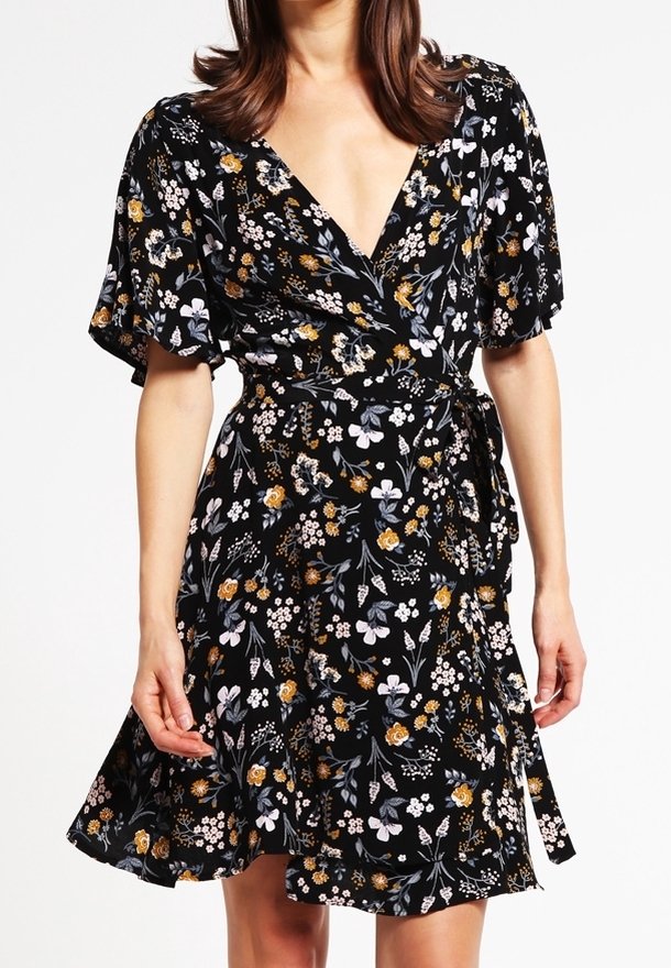Mujer vestida con un vestido envolvente negro con estampado floral, de mangas cortas con volantes y un cinturón atado a la cintura, contra un fondo blanco.