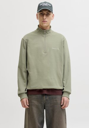 Joven con sudadera verde oliva con cremallera hasta el pecho, vaqueros oscuros, una camisa burdeos debajo y una gorra negra con texto blanco.