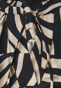 Primo piano di un tessuto con un motivo geometrico astratto in nero e beige, caratterizzato da due fessure verticali e dettagli di cucitura arricciata.