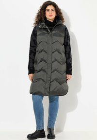 Puffiger, dunkelgrauer, langer Weste mit Kapuze, mit einem Chevron-Muster und Reißverschluss. Kombiniert mit einem schwarzen gemusterten Pullover und blauen Jeans.