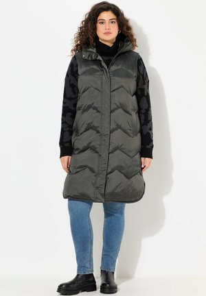 Gilet lungo imbottito, grigio scuro, con cappuccio, caratterizzato da un motivo a zig zag e chiusura con zip. Abbinato a un maglione nero con motivo e jeans blue.