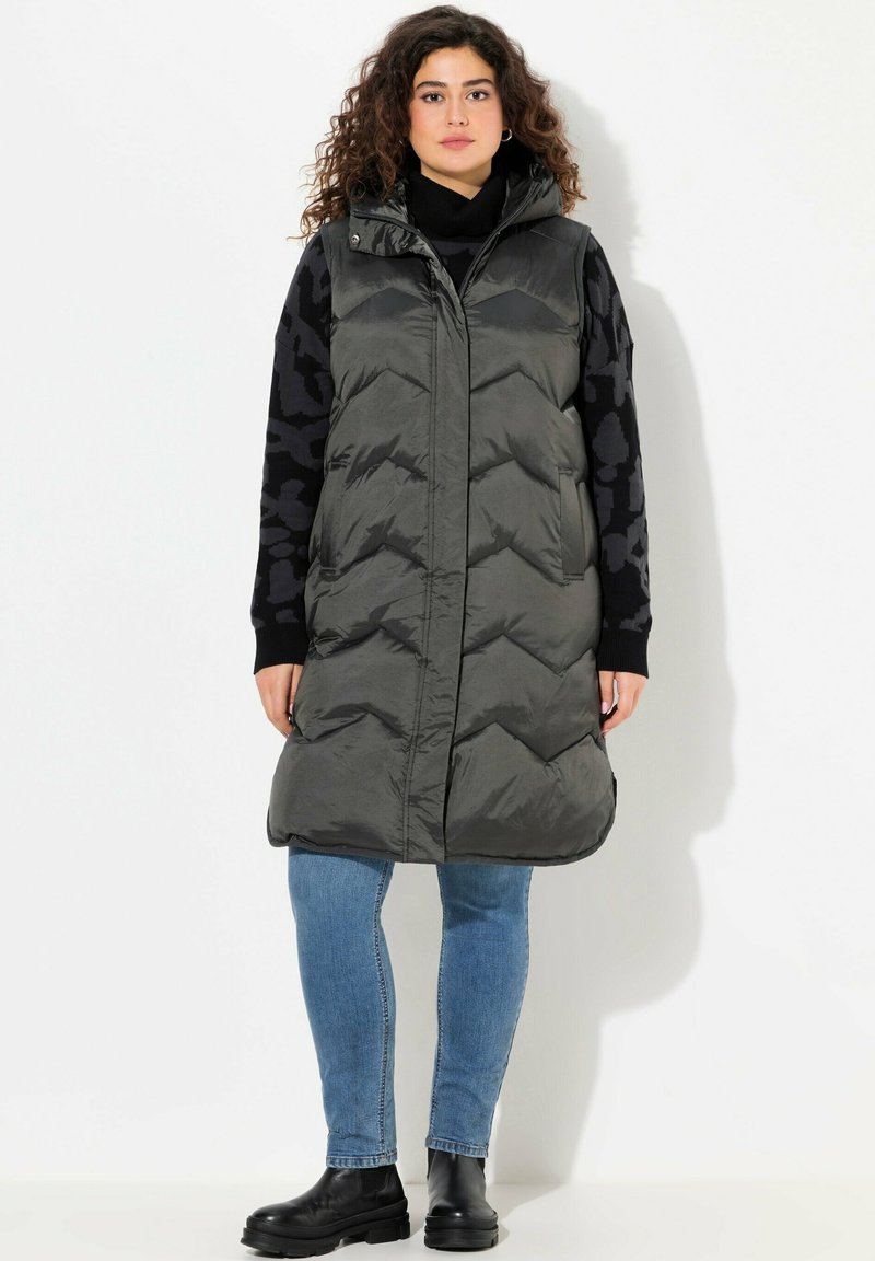 Gilet lungo imbottito, grigio scuro, con cappuccio, caratterizzato da un motivo a zig zag e chiusura con zip. Abbinato a un maglione nero con motivo e jeans blue.