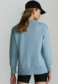 Sweatshirt bleu deschis cu o croială lejeră, cu manșete și tiv ribbate. Asortat cu pantaloni scurți negri, având o vedere din spate și o șapcă neagră.