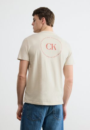 Mladý muž otočený chrbtom, nosiaci béžové tričko Calvin Klein s červeným logom CK a modré džínsy, na jednoduchom bielom pozadí.