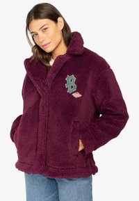 Giacca in pile bordeaux con una texture morbida, vestibilità oversize, collo scialle e toppa ricamata con una "B" verde e dettagli colorati.