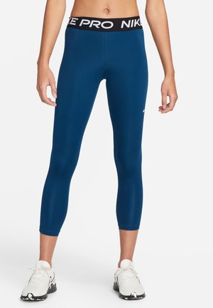 Femme portant un legging court Nike Pro bleu et des chaussures de sport blanches, debout avec les bras détendus le long du corps sur un fond blanc.