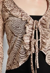 Gros plan d’un chemisier transparent beige et marron à motifs avec des bords volantés noués à l’avant, porté sur un haut noir.