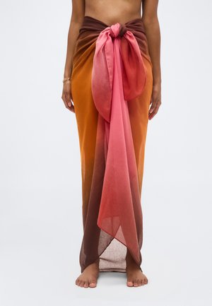 Kvinne iført en gjennomsiktig sarong med oransje til rosa gradient, knyttet i en stor knute i midjen, stående barfot mot en ensfarget bakgrunn.