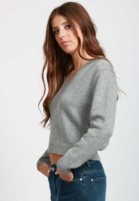Giovane donna con lunghi capelli castani che indossa un maglione grigio a scollo a V con rifinitura di perle e jeans blu, mani nelle tasche, che guarda di lato.