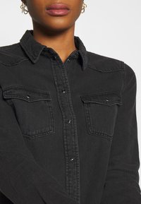 Camicetta in denim nero con colletto a punta, due tasche sul petto e bottoni a pressione neri. Il tessuto ha una leggera texture ed è caratterizzata da una vestibilità aderente.