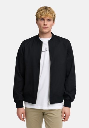 Junger Mann mit kurzen blonden Haaren, trägt eine schwarze Reißverschlussjacke über weißem T-Shirt und beigen Hosen, steht vor schlichtem Hintergrund.