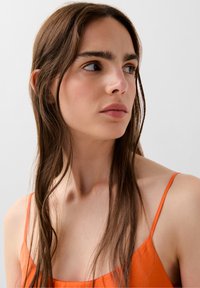 Orange spaghetti-stropkjole med et glat stof. Langt, strait brunt hår delvist rammer ansigtet. Neutral baggrund.
