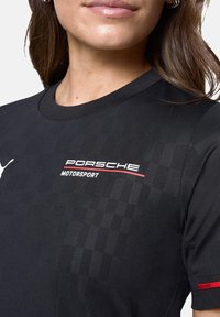 Czarna koszulka sportowa z okrągłym dekoltem, teksturowanym materiałem i logo Porsche Motorsport w białym i czerwonym kolorze z przodu.