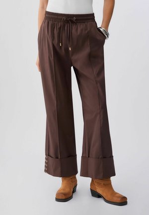 Personne portant un pantalon marron ample avec de larges poignets et des bottines en daim beige sur un fond uni.