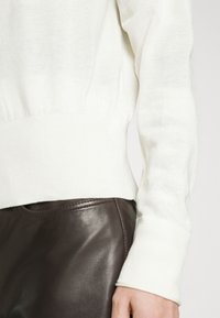 Bílý pletený svetr s žebrovanými manžetami a přiléhavým pasem, doplněný hladkými hnědými koženými kalhotami. Jemný detail loga u lemu.