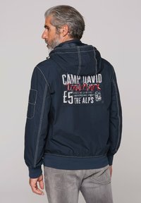 Navy blue bomber jacket with a hood, featuring white stitching and a logo patch on the back. Includes a front pocket and elastic cuffs.