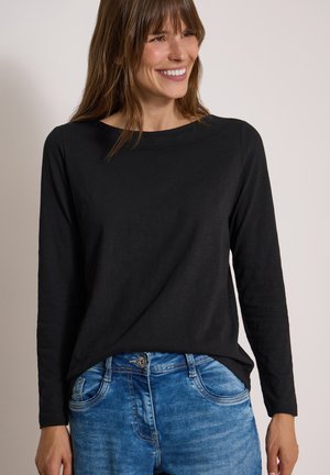Femme souriante portant un haut noir à manches longues et un jean bleu sur un fond clair uni.