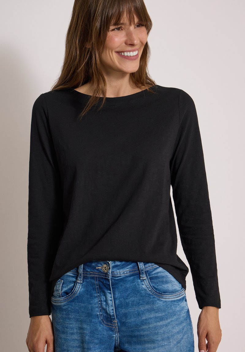 Femme souriante portant un haut noir à manches longues et un jean bleu sur un fond clair uni.