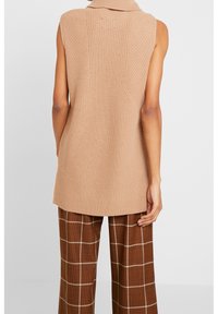 Strikvest i lys beige med høj krave, ærmeløs design, tekstureret sting og en lidt længere ryg. Kombineret med brune ternede bukser.