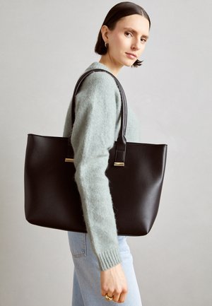 Woman with short dark hair wearing gray sweater and jeans, carrying large black leather tote bag on shoulder against plain background.