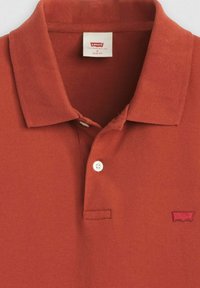 Rustfärgad polo med strukturtyg. Har en traditionell krage, två knappar och en liten broderad logotyp på bröstet.