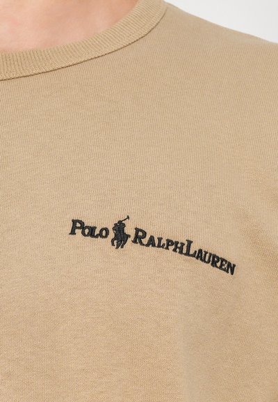 Bēša kreklveida T-krekls ar teksturētu audumu, kuram ir melns izšūts "Polo Ralph Lauren" logo ar ikonisku jātnieka grafiku.