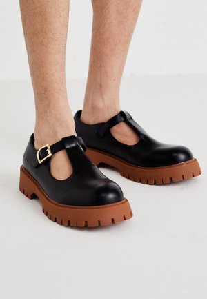 Zwarte leren T-band schoenen met een chunky bruine zool, voorzien van een gespsluiting en een ronde neus. De ribbelstructuur van de zool is zichtbaar.