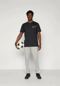 Black short-sleeve t-shirt with a red and green logo on the chest, paired with light gray sweatpants featuring white side stripes, holding a soccer ball.
