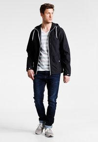 Black lightweight jacket with a hood, front zipper, and white drawstrings, worn over a gray striped t-shirt and blue jeans.