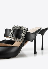 Mule nere in pelle con tacco alto, dotate di una grande fibbia decorata con strass e una superficie liscia. Presentano punta aperta e tacco a stiletto sottile.
