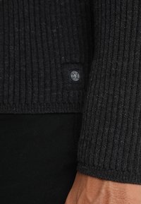 Maglione nero a costine con un design di maglia testurizzata e una piccola etichetta con logo sul lato. Il tessuto appare morbido e caldo.