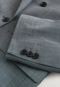 Manche de veste de costume à carreaux gris avec trois boutons noirs pliée sur le corps de la veste assortie avec deux boutons noirs visibles.