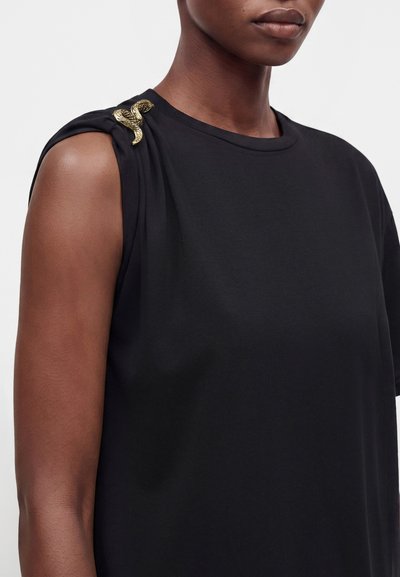 Personne portant un haut noir sans manches avec une broche en forme de serpent dorée sur l'épaule gauche sur un fond uni.