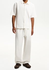 Vit kortärmad skjorta med knappar i texturerat tyg, ihop med löst sittande vita byxor. Bruna träningarskor fulländar outfiten.