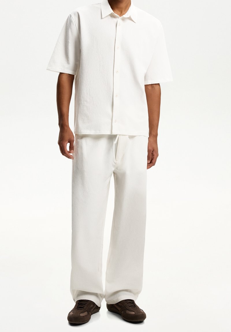 Vit kortärmad skjorta med knappar i texturerat tyg, ihop med löst sittande vita byxor. Bruna träningarskor fulländar outfiten.