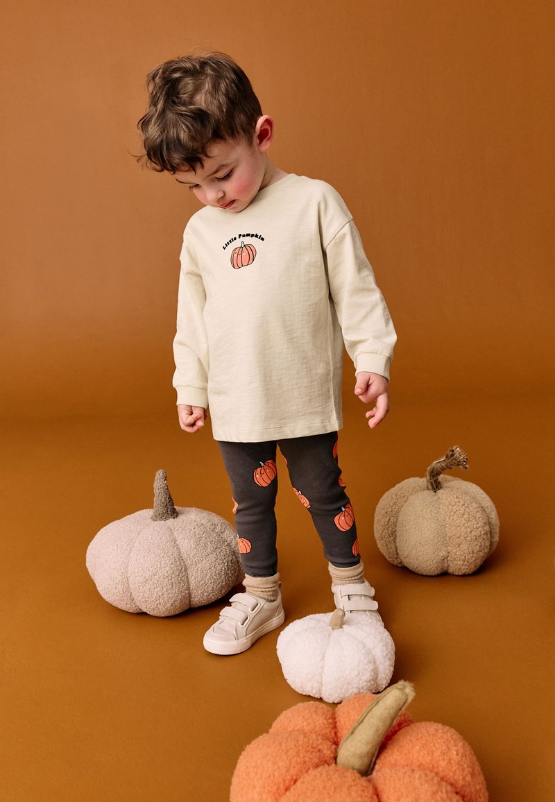 Chemise beige clair à manches longues avec un motif de citrouille, associée à des leggings noirs ornés de motifs de citrouilles répétées, à côté de citrouilles douces et moelleuses.