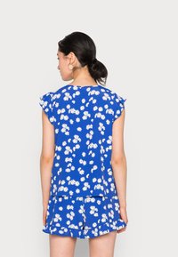 Top floral azul com padrão de margaridas brancas, mangas curtas com fru-fru e um conjunto de calções franconos combinando. Tecido leve, corte descontraído.