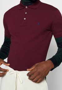 Burgundská polo košile z bavlněné směsi, s klasickým límečkem, zapínáním na tři knoflíky a vyšitým modrým logem. S zelenými kostkovanými rukávy.