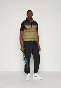 Weste mit Steppung in olivgrünen und schwarzen Abschnitten, getragen über einem schwarzen Hemd. Das Model hält einen Schläger, kombiniert mit schwarzen Hosen und weißen Sneakers.