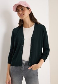 Donkergroene cardigan met een V-hals, knoopsluiting aan de voorkant en geribbelde manchetten, gecombineerd met een lichte top en grijze jeans; model draagt een roze pet.