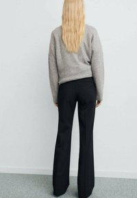 Maglione grigio lavorato a maglia con maniche lunghe, abbinato a pantaloni neri a vita alta, visto da dietro in un ambiente minimalista.