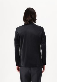 Chaqueta de traje negra hecha de tela suave; presenta solapas muestradas, abertura trasera y acentos de puños abotonados. Vista desde atrás.
